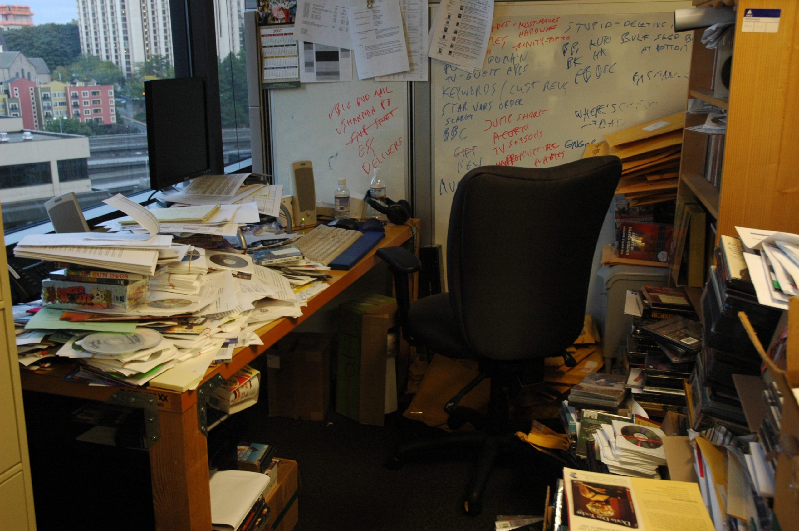A desk and a chair in an office, with disorganized piles of papers everywhere.