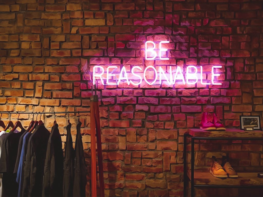 a neon sign saying "Be Reasonable" against a brick wall backdrop